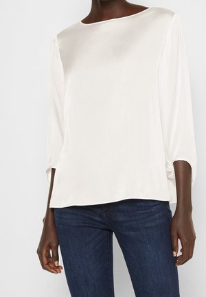 Weiße Satinbluse mit einem Boots-Ausschnitt und 3/4-Ärmeln. Verfügt über eine lockere Passform und einen dezenten Glanz. Kombiniert mit dunkelblauen Jeans.