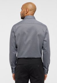 Camicia a maniche lunghe di colore blu scuro con un motivo geometrico, dotata di un colletto classico e polsini con bottoni. Abbinata a pantaloni neri.