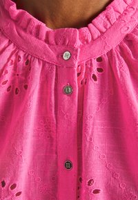 Blusa de algodão rosa com um decote franzido, intrincada bordadura floral e botões metálicos na frente. A textura parece leve e arejada.