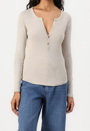 Femme portant un haut beige côtelé à manches longues avec un col en V court boutonné, associé à un jean denim bleu moyen.