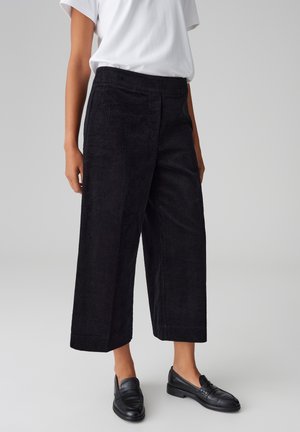 Mujer con pantalones cortos de pana negra de pierna ancha, camiseta blanca y mocasines de cuero negros, de pie sobre un fondo gris liso.