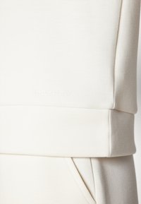 Lys beige sweatshirt med en myk tekstur, ribbestrikket midjebånd og subtilt preget logo. Designet med sidelommer og en figursydd fasong.