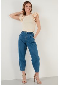 Světle béžový top s volánovými rukávy, zkombinovaný s modrými džíny s vysokým pasem. Boty jsou béžové lodičky. Čisté pozadí, model pózuje s jednou rukou zdviženou.