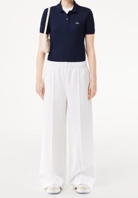 Polo-shirt bleu marine avec col, pantalon blanc à taille élastique, sac beige et baskets claires avec accents bleus. Texture lisse, design décontracté.