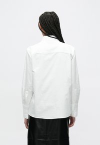 Bijela košulja s dugim rukavima s vrhunskim ovratnikom, manžetnama na kopčanje i ravnim stražnjim dizajnom, uparena s crnom kožnom suknjom. Glatka tekstura.