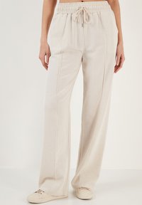 Beige wijde benen broek van zacht materiaal met een elastische tailleband en een trekkoord. Heeft voornaadjes en een losse pasvorm voor comfort.