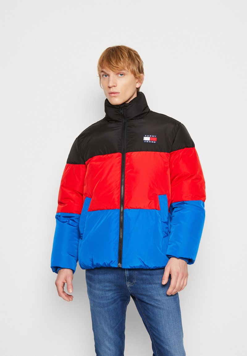 Giacca imbottita con parte superiore nera, sezione centrale rossa e parte inferiore blu. Collo alto, zip frontale e patch del logo sul petto.