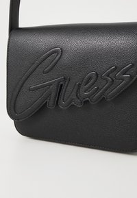 Črna umetna usnjena torbica čez ramo z izbočenim logotipom "Guess", gladko teksturo in zapiranjem s preklopom ter enojnim trakom.