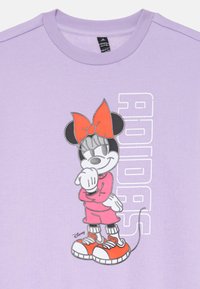 T-shirt viola con un contorno grigio di un personaggio dei cartoni animati in un abito rosa con un fiocco e scarpe arancioni; il testo "ADIDAS" è visibile.