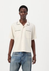 Short-sleeve, cream button-up shirt with black piping, two front pockets, and a relaxed fit. Worn with light blue denim jeans.