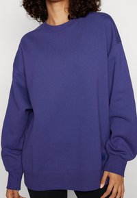 Sudadera púrpura hecha de material suave, con cuello redondo, mangas largas, corte holgado y puños y dobladillo acanalados, con un logo mínimo.