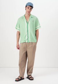 Green short-sleeve shirt with geometric pattern and buttons, paired with light beige pants and black sandals. Light blue cap accessory included.