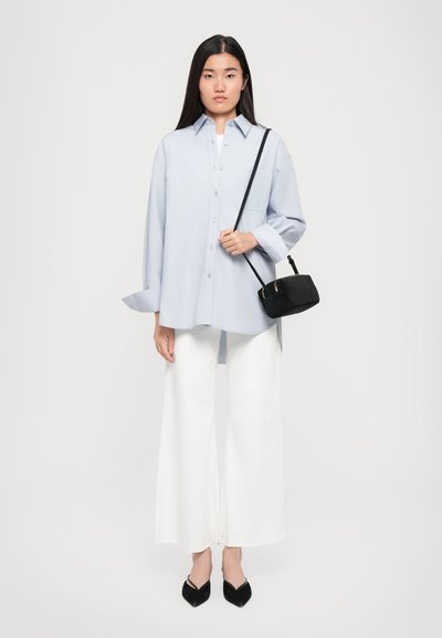 Camicia azzurro chiaro con maniche lunghe, abbinata a pantaloni bianchi larghi e una piccola borsa nera; modello in piedi contro uno sfondo neutro.