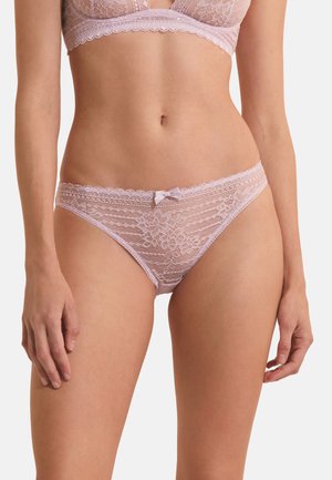 Femme portant un soutien-gorge en dentelle rose clair et une culotte en dentelle assortie avec un motif floral et un petit nœud sur la taille de la culotte.