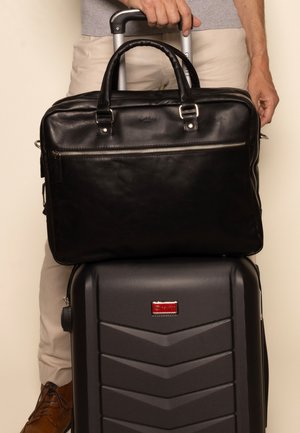 Black leather briefcase with dual handles, silver zipper, placed atop a black hard-shell suitcase with textured chevron patterns and a red logo.
