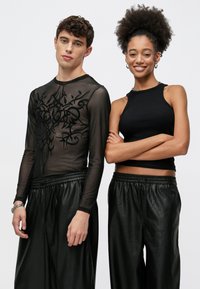 Μαύρο μακρυμάνικο τοπ από δίχτυ με περίπλοκο σχέδιο, σε συνδυασμό με φαρδιά μαύρα παντελόνια από ψεύτικο δέρμα· το μοντέλο αριστερά φοράει αμάνικο τοπ.