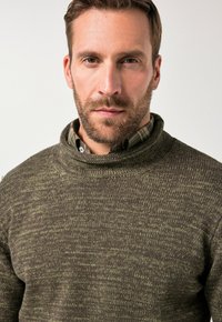 Suéter de punto grueso y verde oscuro con un cuello alto y doblado y una textura sutil. Debajo, se asoma una camisa a cuadros verde y negra.