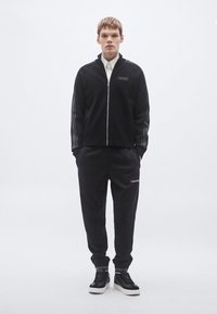 Casaco preto com fecho de correr, gola alta, riscas laterais e aplicação com logótipo; combinado com calças de fato de treino pretas com um logótipo perto da bainha.