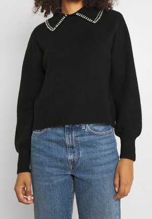 Pull noir à manches longues avec col orné de perles porté avec un jean bleu taille haute sur une personne aux cheveux bouclés.