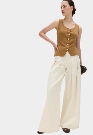 Mujer con top marrón sin mangas y abotonado, pantalones anchos color crema, zapatos negros y sosteniendo un pequeño clutch oscuro.