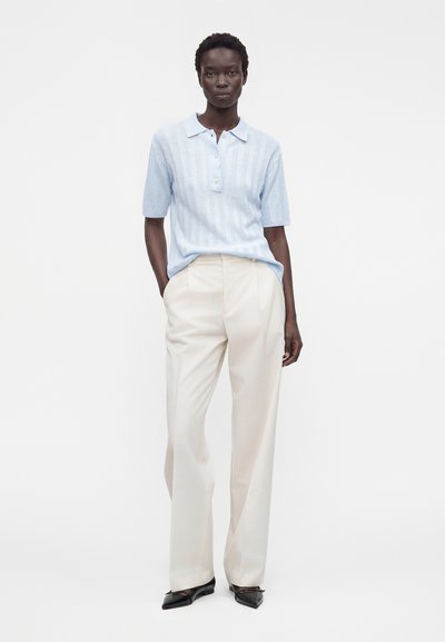 Personne debout portant un polo en maille bleu clair à manches courtes, un pantalon crème à jambes larges et des ballerines pointues noires sur fond blanc.