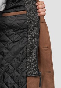 Hnědý vnější kryt s prošívanou černou podšívkou a strukturovaným šedým interiérem. Zahrnuje zip a vnitřní kapsu. Jednoduchý design s čistými liniemi.