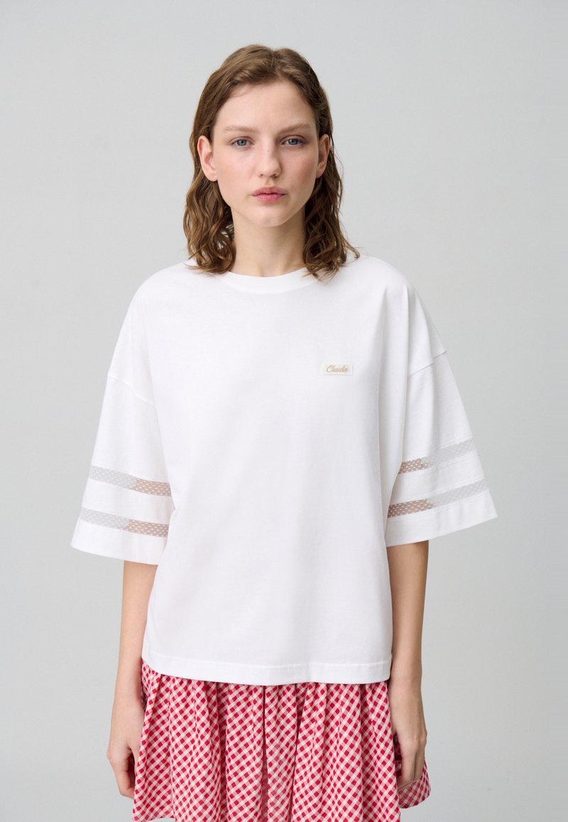 T-shirt bianco oversize con maniche corte, caratterizzato da dettagli a strisce in rete e una piccola etichetta con logo. Tessuto morbido con vestibilità rilassata.