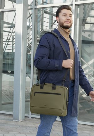 Mann in blauem Mantel und beigem Rollkragenpullover trägt eine olivgrüne Umhängetasche und geht vor einem modernen Glasgebäude.