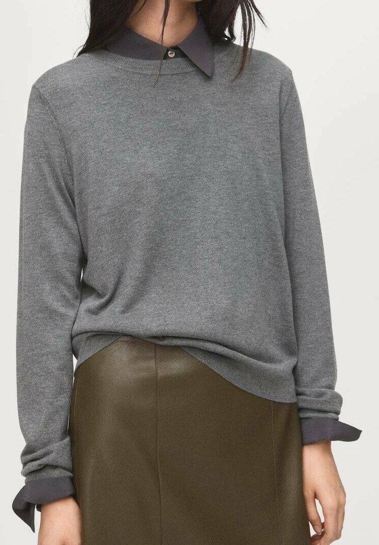 Femme portant un pull à manches longues gris sur une chemise à col foncé et une jupe en cuir marron, debout devant un fond clair uni.