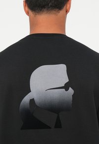 Maglietta nera con una grafica a silhouette grigia sfumata di una persona che indossa occhiali da sole sulla schiena. Design semplice con scollatura a girocollo.