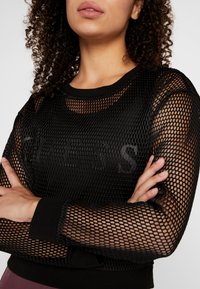 Czarna siateczkowa bluzka z długim rękawem i okrągłym dekoltem, z nadrukowanym logo na piersi, żebrowanymi mankietami i luźnym krojem.