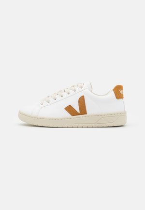 Bílá nízká teniska s béžovou podrážkou, béžovými tkaničkami a hnědým semišovým logem ve tvaru "V" a patní záložkou na jednobílém pozadí.