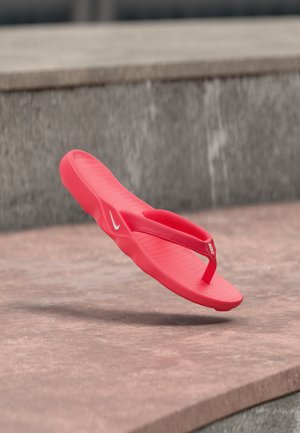 Rød Nike flip-flop sandalen hænger over en stenet overflade med en grå tekstureret væg som baggrund.