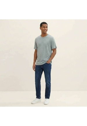 Homme en chemise grise à manches courtes, jean bleu foncé et baskets blanches, debout avec une main dans la poche, souriant et regardant sur le côté.