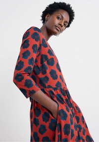 Rövid göndör hajú nő, aki vörös ruhát visel, nagy sötétkék virágmintákkal, kezét a zsebében tartja, egyszínű világos háttér előtt.