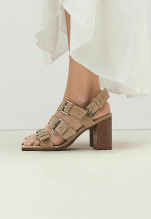 Žena nosi béžové semišové sandále na hrubom podpätku s tromi nastaviteľnými remienkami so sponami a bielu splývavú sukňu.