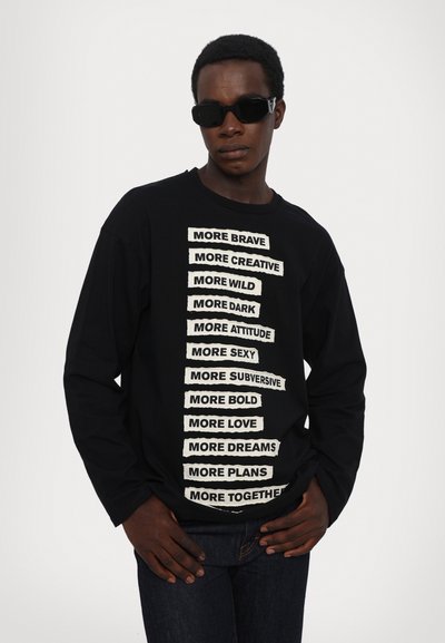 Black long-sleeve t-shirt with white text bands featuring various phrases down the center. Model wearing black sunglasses and denim jeans.