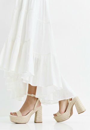 Jambes en talons compensés tissés beige avec des brides à la cheville, marchant sous une longue jupe blanche fluide à volants.