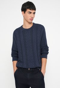 Granatowy sweter dziany z wzorem warkocza, okrągłym dekoltem i długimi rękawami. Do tego ciemne spodnie, ręce w kieszeniach.