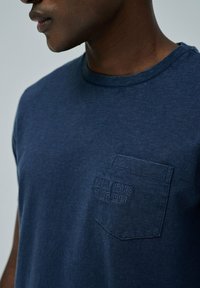 Camiseta de algodón azul marino de manga corta, con un bolsillo en el pecho y texto en relieve que dice "Salsa Jeans Since 1994." Textura suave.