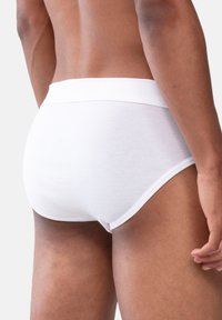 Vue arrière d'une personne portant des slips blancs simples, montrant la taille, les hanches, le haut des cuisses et une main détendue sur le côté.