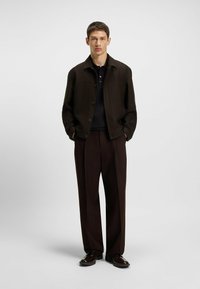 Giacca marrone scuro con chiusura a bottone, indossata su una polo nera, abbinata a pantaloni larghi marrone scuro e scarpe lucide.