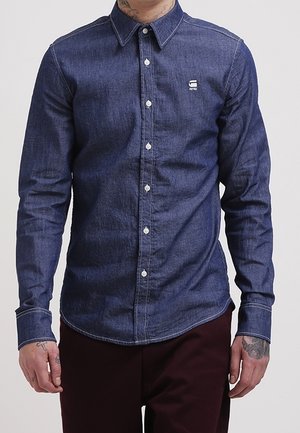 Mand iført en tætsiddende, knappet mørkeblå denimskjorte med hvide knapper og burgunderbukser, synlige tatoverede hænder og nakke.