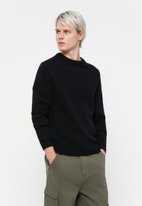 Pulover negru tricotat cu guler rotund cu nervuri, mâneci lungi și o croială lejeră, asortat cu pantaloni cargo de culoare verde măsliniu.