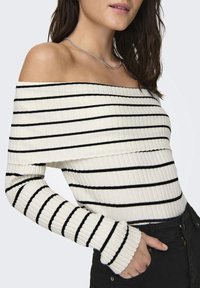 Biały prążkowany sweter odkrywający ramiona z poziomymi czarnymi paskami, z długimi rękawami zakończonymi dopasowanymi mankietami. Stylizowany z czarnymi spodniami.