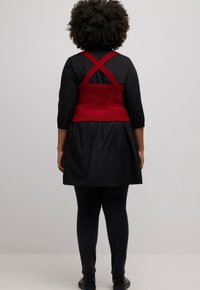 Red cross-back top over a black dress with three-quarter sleeves, paired with dark leggings and black shoes. Smooth, structured fabric.