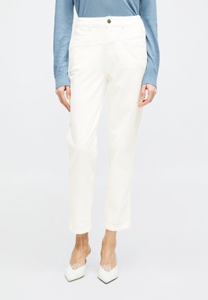 Kvinde iført hvide højtaljede bukser med lige ben, kombineret med hvide spidse hæle og en lyseblå langærmet top.