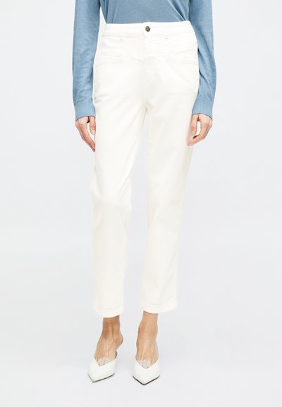Femme portant un pantalon blanc taille haute à jambes droites associé à des talons blancs à bout pointu et un haut bleu clair à manches longues.