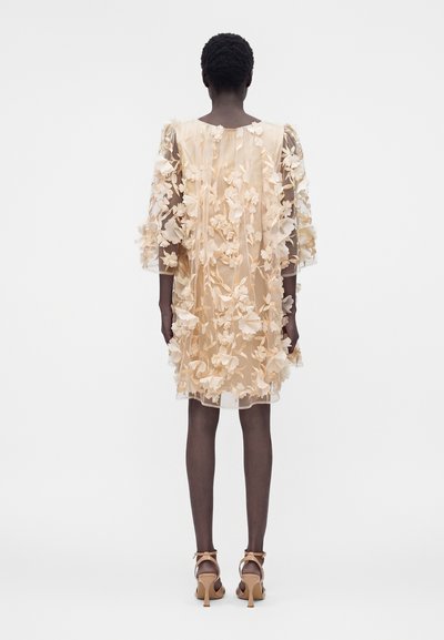 Γυναίκα με διάφανο μπεζ φόρεμα μέχρι το γόνατο με 3D λουλουδένιες εφαρμογές και μανίκια μέτριου μήκους, σε συνδυασμό με μπεζ σανδάλια με ψηλό τακούνι, εμφανίζεται από πίσω.