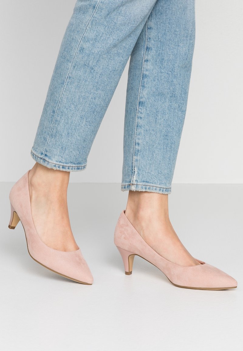Pieds portant des escarpins rose clair en daim à talon bas et bout pointu, associés à un jean bleu clair court sur un fond uni.